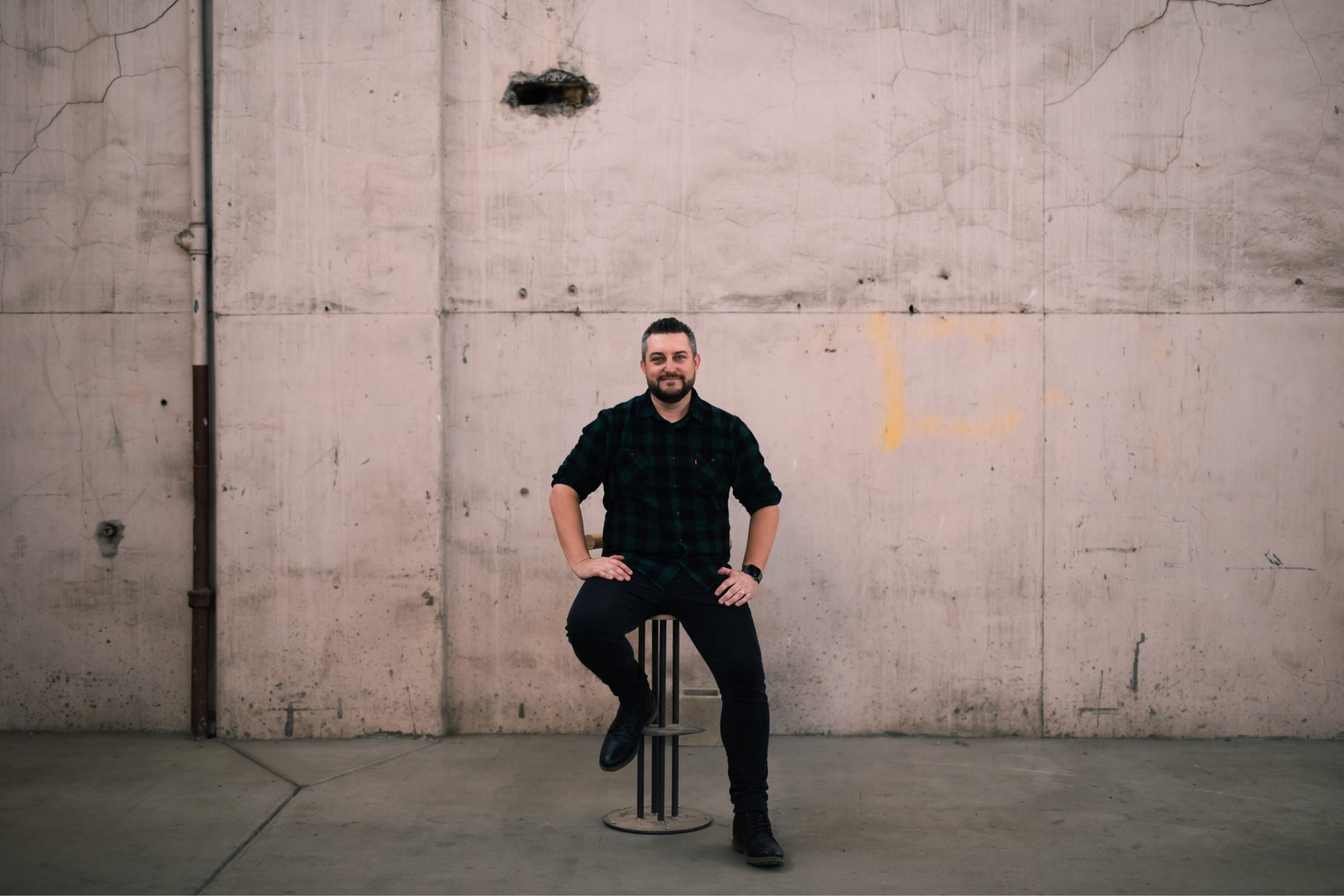 Joshua Brown seated on a stool in front of a pale concrete wall.
